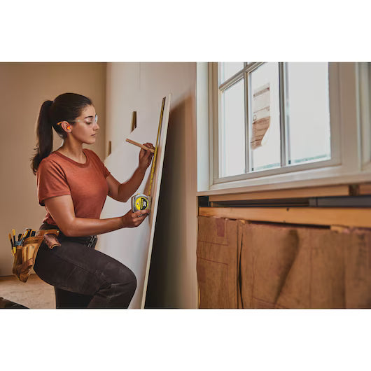 Kneeling indoors by a window, a woman wearing a tool belt measures and marks drywall with the STANLEY 25 ft PowerLock® Tape Measure and pencil, surrounded by sturdy construction tools on her belt.