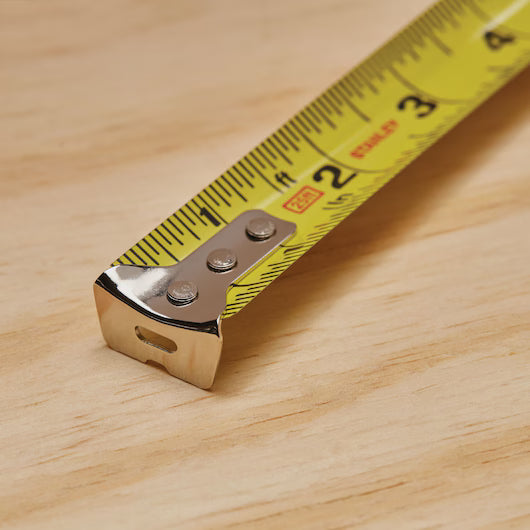 Close-up of a STANLEY 25 ft PowerLock® Tape Measure extended on a light wooden surface, showing inch and centimeter markings with its metal end in the foreground.