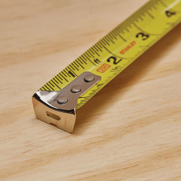 Close-up of a STANLEY 25 ft PowerLock® Tape Measure extended on a light wooden surface, showing inch and centimeter markings with its metal end in the foreground.