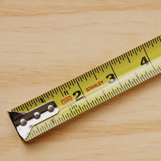 A close-up of a durable 25 ft PowerLock® Tape Measure by STANLEY, extended to 3 inches on a light wooden surface, with clear numbers, markings, and the STANLEY brand name visible.