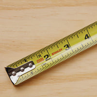 A close-up of a durable 25 ft PowerLock® Tape Measure by STANLEY, extended to 3 inches on a light wooden surface, with clear numbers, markings, and the STANLEY brand name visible.