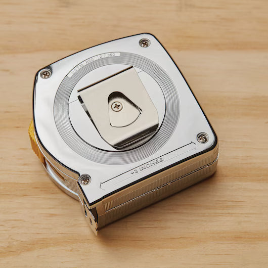 A STANLEY 25 ft PowerLock® Tape Measure with a metal belt clip sits on a light wooden surface. The tape is partially retracted, and its square, rounded-edge casing reflects the tool’s durability.