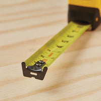 A close-up of the STANLEY® FATMAX® Classic 25 ft Tape Measure, extended over a wooden surface with clear inch and fraction markings, features the metal end hook in the foreground.