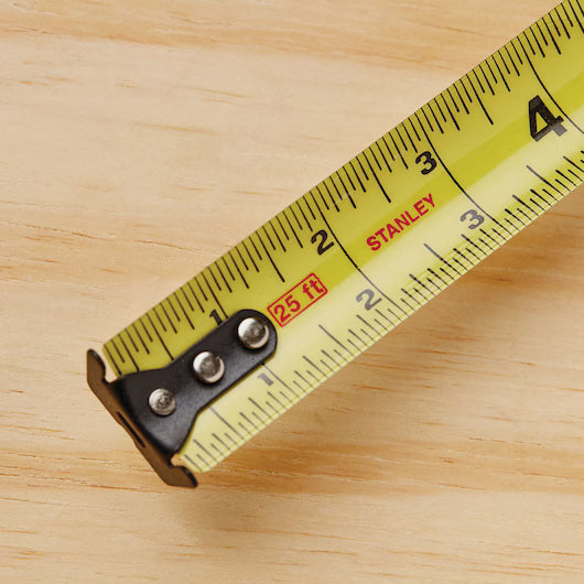 A close-up of a yellow 25 ft STANLEY® FATMAX® Classic Tape Measure extended to about 4 inches on a light wood surface, showing black and red inch and foot markings.