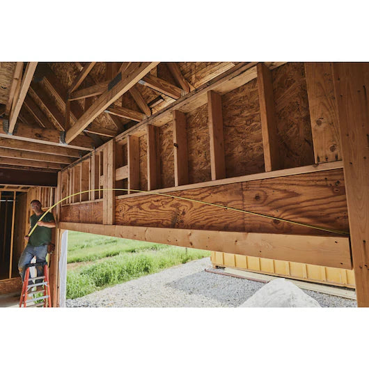 A person on a ladder inside a wooden house under construction runs yellow electrical wire through wall studs, with a 30 ft STANLEY® FATMAX® Classic Tape Measure nearby. Green grass and gravel can be seen outside.