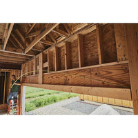 A person on a ladder inside a wooden house under construction runs yellow electrical wire through wall studs, with a 30 ft STANLEY® FATMAX® Classic Tape Measure nearby. Green grass and gravel can be seen outside.