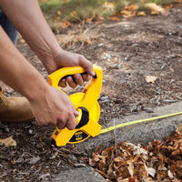 A person uses the STANLEY 100 ft Fiberglass Long Tape to measure distance outdoors on soil with leaves and grass, extending the yellow measuring tool along the ground.