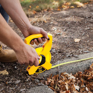 A person uses the STANLEY 100 ft Fiberglass Long Tape to measure distance outdoors on soil with leaves and grass, extending the yellow measuring tool along the ground.