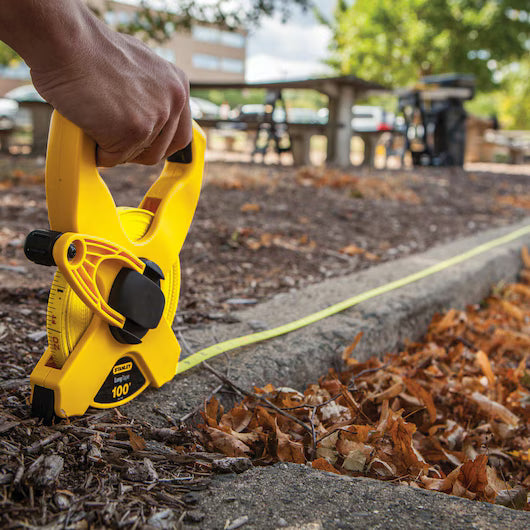 A person uses a STANLEY 100 ft Fiberglass Long Tape to measure distance along a leaf-covered sidewalk, with picnic tables and trees in the background.