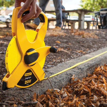 A person measures curb distance with a STANLEY 1/2 in x 200 ft Open Reel Fiberglass Long Tape; fallen leaves and an outdoor table are visible in the background.
