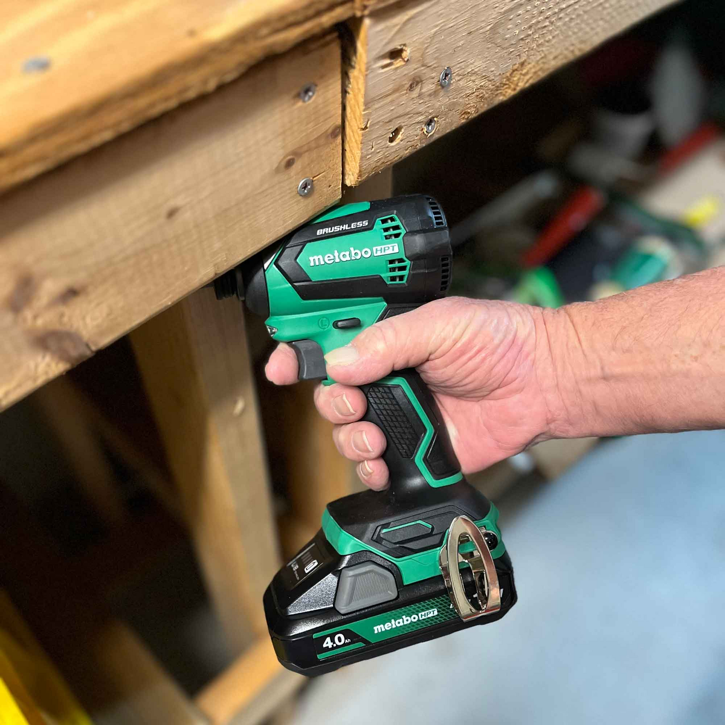 A person uses a Go Build Metabo HPT 378681M BSL1840M drill with an 18V 4.0Ah Lithium Ion Battery and fuel indicator to drive a screw into wood, with a workshop workbench and tools visible in the background.