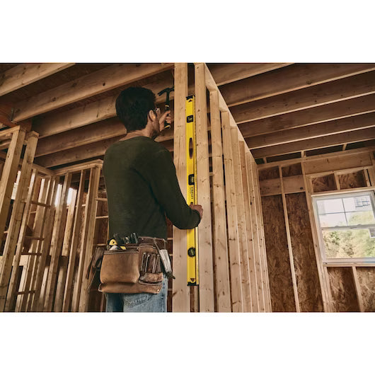 A worker wearing a tool belt uses the STANLEY 48 in I-Beam 180 Level to check the vertical alignment of a wooden frame inside an unfinished building during construction.