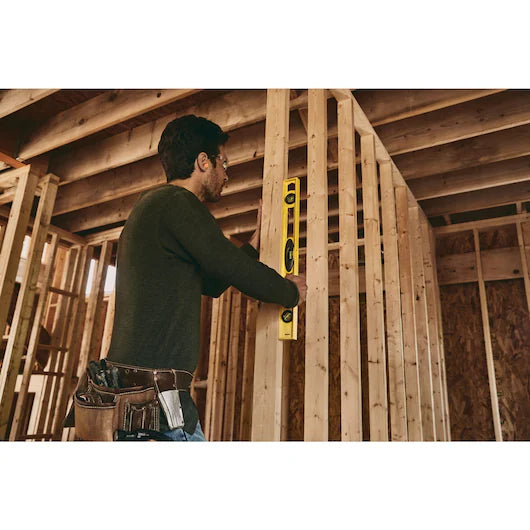 A person wearing a tool belt uses a STANLEY 24 in High-Impact ABS Level to check the alignment of wooden framing amid exposed beams and unfinished walls in a building under construction.