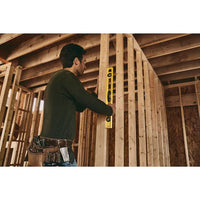 A person wearing a tool belt uses a STANLEY 24 in High-Impact ABS Level to check the alignment of wooden framing amid exposed beams and unfinished walls in a building under construction.