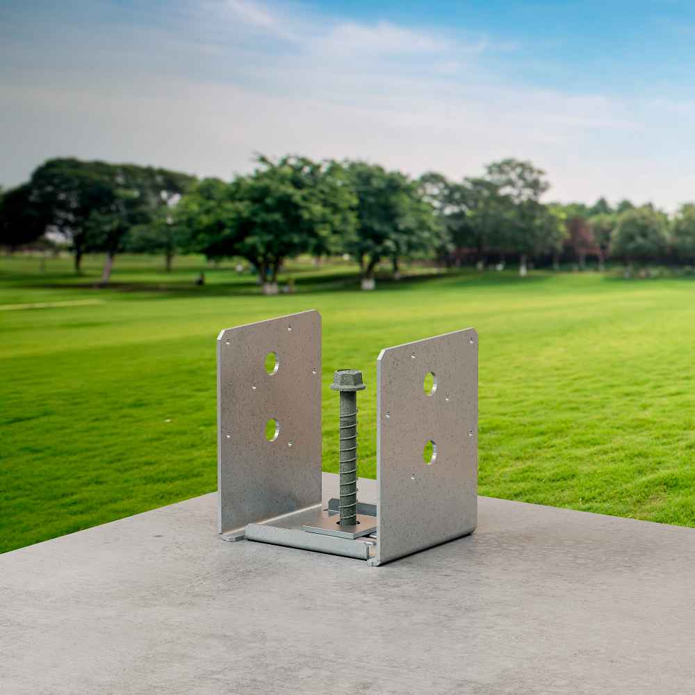 The Simpson Strong-Tie ABU ZMAX® 4-in x 4-in post base bracket, ideal for connecting wood posts to concrete in high-capacity uses, features four holes and a central bolt. It is shown on concrete near a grassy field with trees under a blue sky.