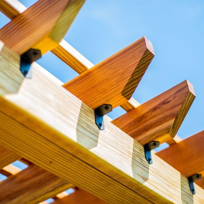 Close-up of a wooden pergola with slatted rafters against a bright blue sky. Supported by the SIMPSON APA21 APA 90° Angle from Simpson Strong-Tie, the beams highlight a sturdy yet aesthetic construction, ideal for outdoor projects showcasing natural wood textures.