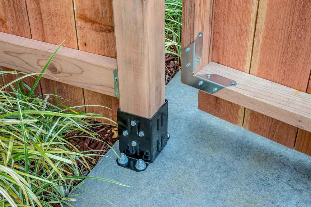 A wooden fence post is anchored to concrete with a Simpson Strong-Tie E-Z Base Black Powder-Coated Post Base for 4x4 lumber and bolts, supporting a wooden gate with metal braces, surrounded by green plants.