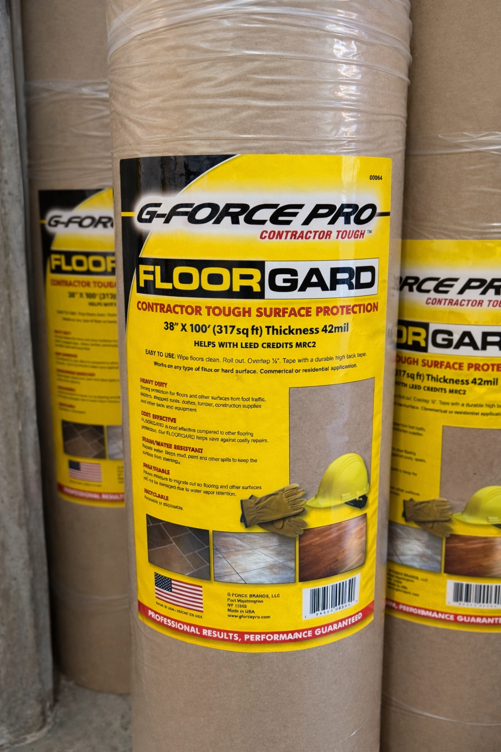 A close-up of a G FORCE 00064 FLOORGUARD 38" x 100' heavy duty extra thick paper roll with a yellow label showing specs and safety info, standing upright among other construction floor covering rolls in the background.