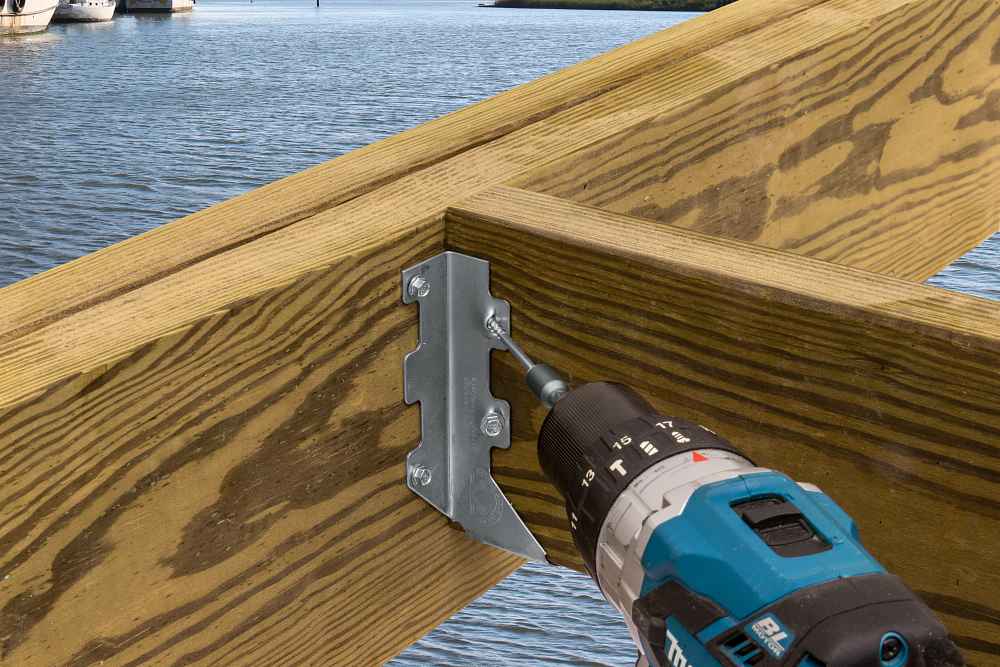 A cordless power drill is being used to secure a Simpson Strong-Tie ZMAX® LUS26Z Single 18-Gauge Face Mount Joist Hanger (2x6) to a wooden beam frame near water, with boats and the opposite shore in the background.