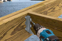 A cordless power drill is being used to secure a Simpson Strong-Tie ZMAX® LUS26Z Single 18-Gauge Face Mount Joist Hanger (2x6) to a wooden beam frame near water, with boats and the opposite shore in the background.