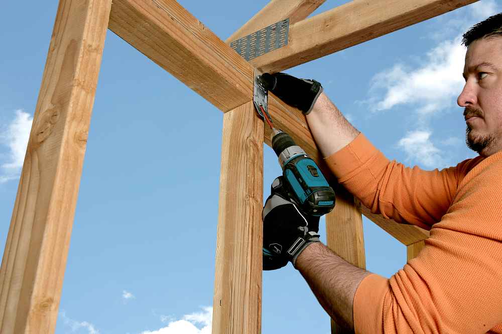 Wearing an orange shirt and gloves, a person uses a power drill to install Simpson Strong-Tie Strong-Drive SDWC TRUSS Screws (6" T30 50CT) for stud-to-plate connection on wood framing under a blue, cloudy sky.