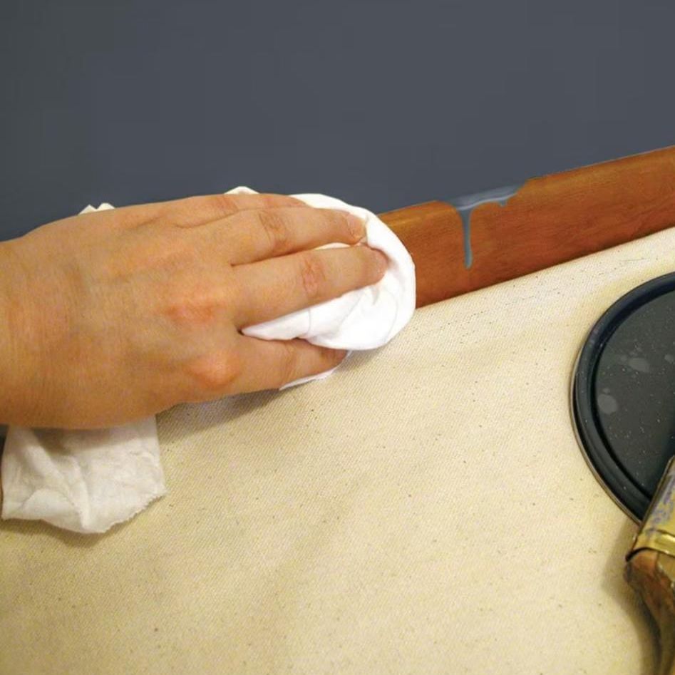 Someone uses PAINT FORCE 36100 bright white knit rags to clean a brown wooden surface with gray paint. Nearby, a black circular object and tin can are partially visible on the beige fabric, making these absorbent 100% cotton cloths ideal for painting projects.