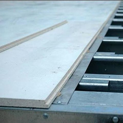 A close-up shows the installation of a Go Build DragonBoard MGO Subfloor Panel, revealing the metal support structure underneath. The 3/4 x 4 x 8 gray fire-resistant panel has a smooth texture, emphasizing where it meets the support grid.