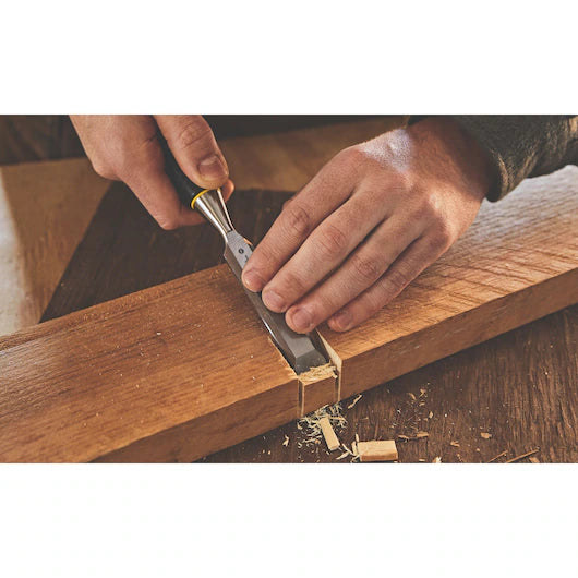 Using the STANLEY® WOOD CHISEL 3PC. SET (13, 19, 25mm), a person carves a rectangular notch into wood, scattering shavings across the surface.