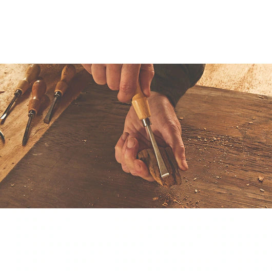 A person carves wood by hand with a STANLEY 6 PC WOOD CARVING TOOL SET, while other chisels and gouges from the set lie nearby, ready for different woodworking projects.