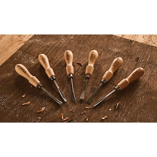 The STANLEY 6 PC WOOD CARVING TOOL SET features six wood chisels and gouges with wooden handles, arranged in a fan on a wooden surface with wood shavings—perfect for all your woodworking projects.