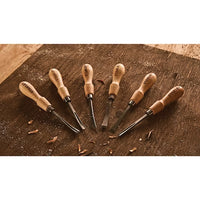 The STANLEY 6 PC WOOD CARVING TOOL SET features six wood chisels and gouges with wooden handles, arranged in a fan on a wooden surface with wood shavings—perfect for all your woodworking projects.