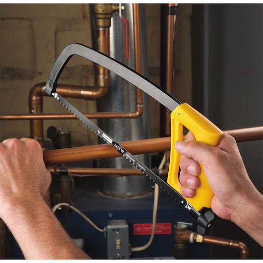 A person uses the STANLEY® D-Shape Hacksaw, featuring a yellow handle and durable steel frame, to cut a copper pipe amid plumbing equipment—demonstrating precision and reliable performance from STANLEY.