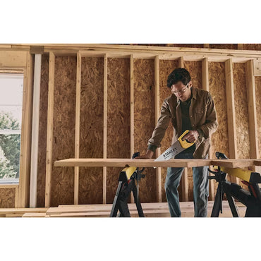 A man uses the STANLEY STHT20348 15 in TradeCut™ Panel Saw to cut a wooden plank supported by two sawhorses in a partially constructed room with exposed wooden framing.