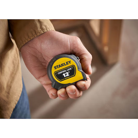 A person holds a STANLEY 12 ft. CONTROL-LOCK™ Tape Measure in their hand, with a blurred indoor background.
