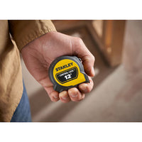 A person holds a STANLEY 12 ft. CONTROL-LOCK™ Tape Measure in their hand, with a blurred indoor background.