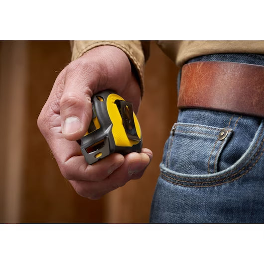 A person in jeans and a brown belt holds the STANLEY 12 ft. CONTROL-LOCK™ Tape Measure, yellow and black, in their right hand with the tape resting against their fingers.