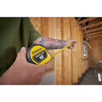 A person with a tattooed arm uses the STANLEY 12 ft. CONTROL-LOCK™ Tape Measure to check the length of a wooden wall frame inside a partially constructed building.