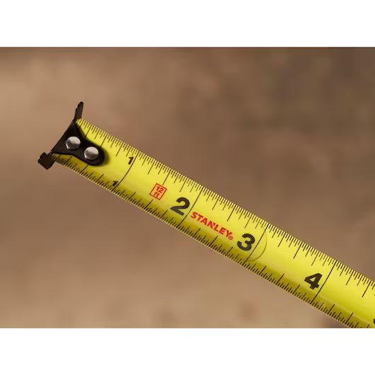 A close-up of the 12 ft. CONTROL-LOCK™ Tape Measure by STANLEY, extended to about 4 inches, shows clear black numbers and markings on its yellow blade against a blurred brown background.