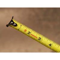 A close-up of the 12 ft. CONTROL-LOCK™ Tape Measure by STANLEY, extended to about 4 inches, shows clear black numbers and markings on its yellow blade against a blurred brown background.