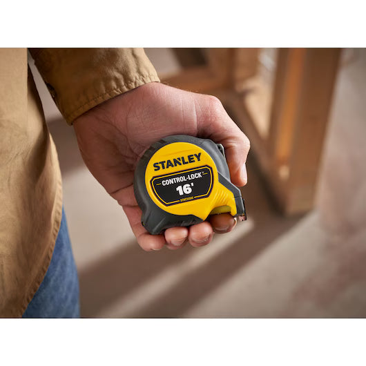 A person holds a STANLEY 16 ft. CONTROL-LOCK™ Tape Measure in their right hand, with a blurred indoor construction setting in the background.