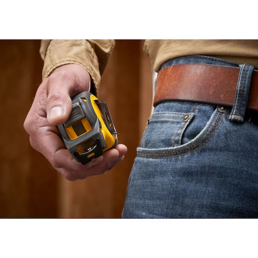 A person in a brown shirt and blue jeans holds a STANLEY 16 ft. CONTROL-LOCK™ Tape Measure in their right hand, standing indoors against a blurred wooden background.