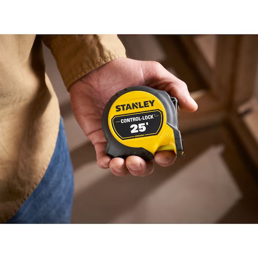 A person holds a STANLEY 25 ft. CONTROL-LOCK™ Tape Measure, its yellow and black design standing out against a blurred indoor background.
