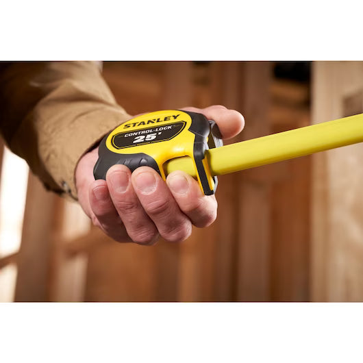 A hand holds a STANLEY 25 ft. CONTROL-LOCK™ Tape Measure, extending it in a room with unfinished wooden walls.