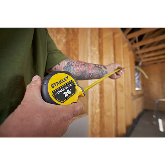 A person with a tattooed arm uses a STANLEY 25 ft. CONTROL-LOCK™ Tape Measure to measure inside a wooden framed room under construction.