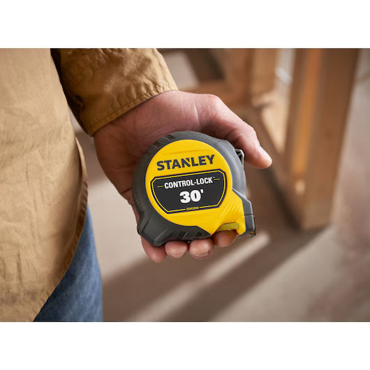 A person in a brown jacket holds a STANLEY 30 ft. CONTROL-LOCK™ Tape Measure, with an indoor construction site blurred in the background.