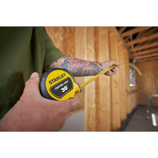A person with a tattooed arm uses a STANLEY 30 ft. CONTROL-LOCK™ Tape Measure to measure distance in a wooden-framed room under construction.