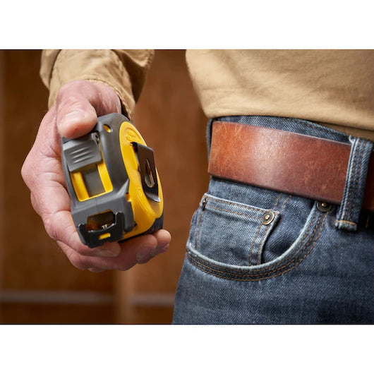 A person wearing blue jeans and a brown belt holds a STANLEY 30 ft. CONTROL-LOCK™ Tape Measure in their right hand, standing indoors against a wooden background.