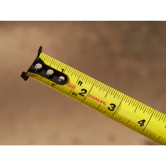A close-up of the end of a STANLEY 30 ft. CONTROL-LOCK™ Tape Measure, extended to nearly 4 inches, shows clear black markings against a blurred brown background.