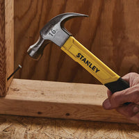 A hand grips a STANLEY 16 OZ CURVE CLAW FIBERGLASS HAMMER, driving a nail into a wooden board with a wooden background.