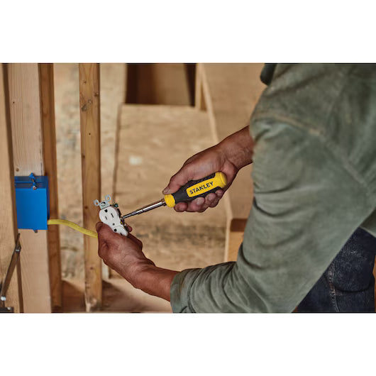A person uses a STANLEY® QUICK CHANGE INTERCHANGEABLE SCREWDRIVER 6-IN-1 to install or adjust an electrical outlet in a wall frame with exposed wooden studs and wires.